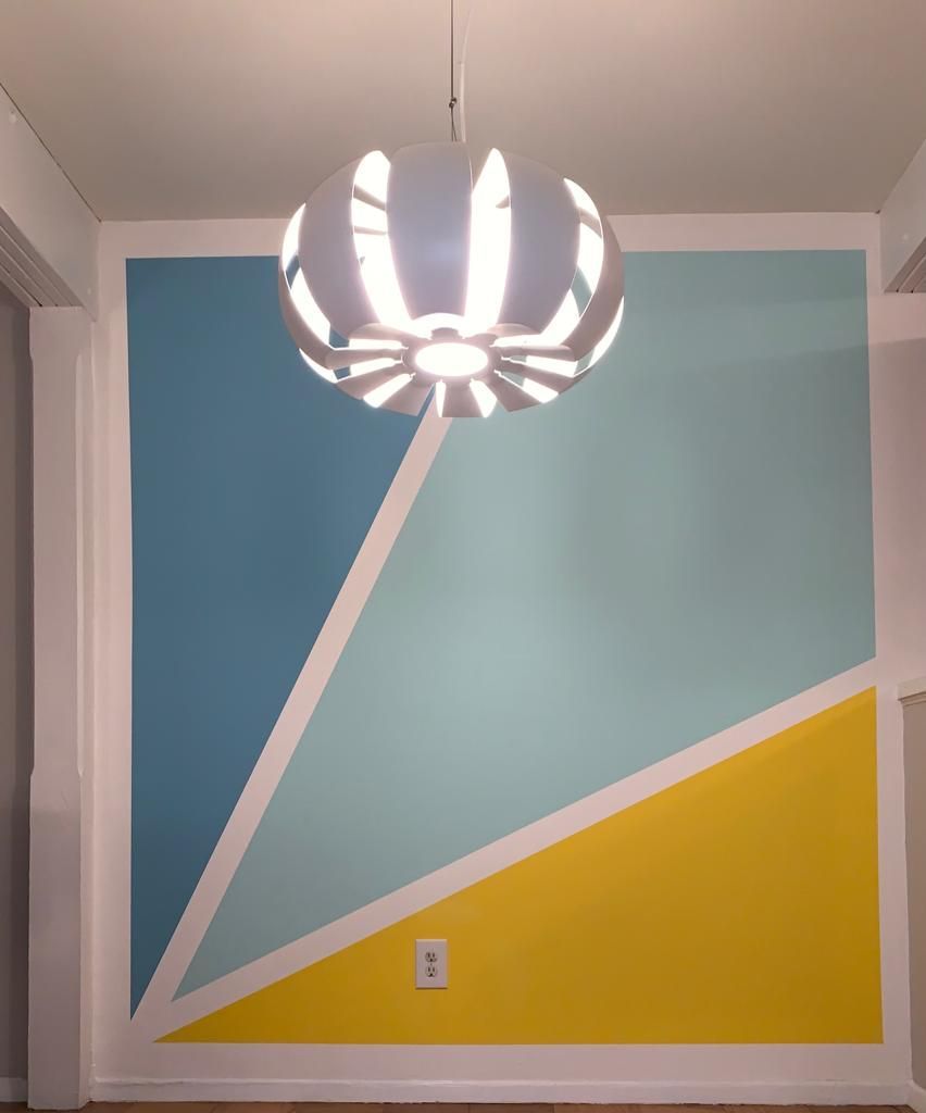 Person painting a wall white with a brush. Paint cans, tray, brushes, and color swatches on the plastic-covered floor.