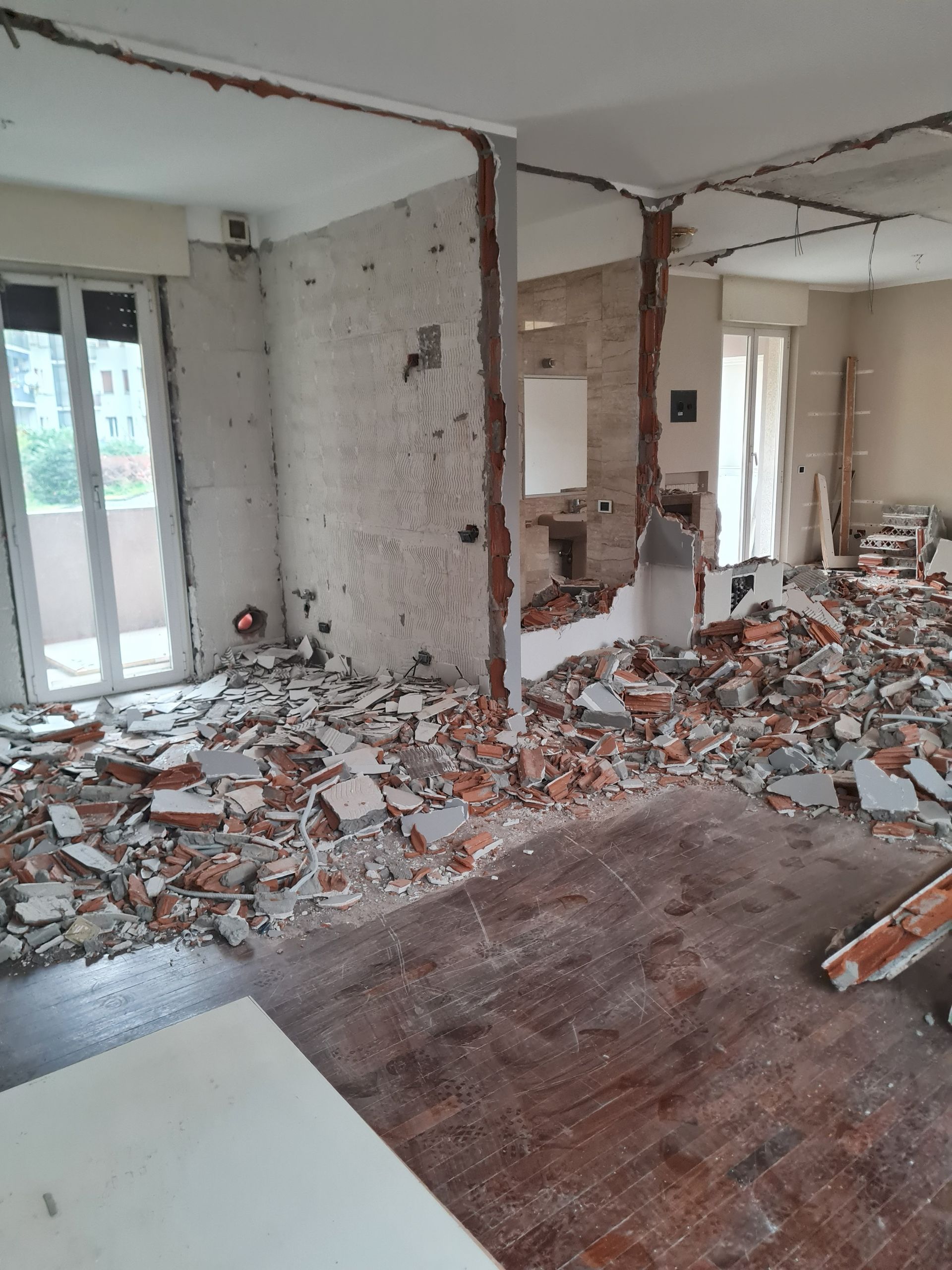 Interno in fase di demolizione, mattoni e detriti sul pavimento, pareti parzialmente rimosse, finestre visibili.