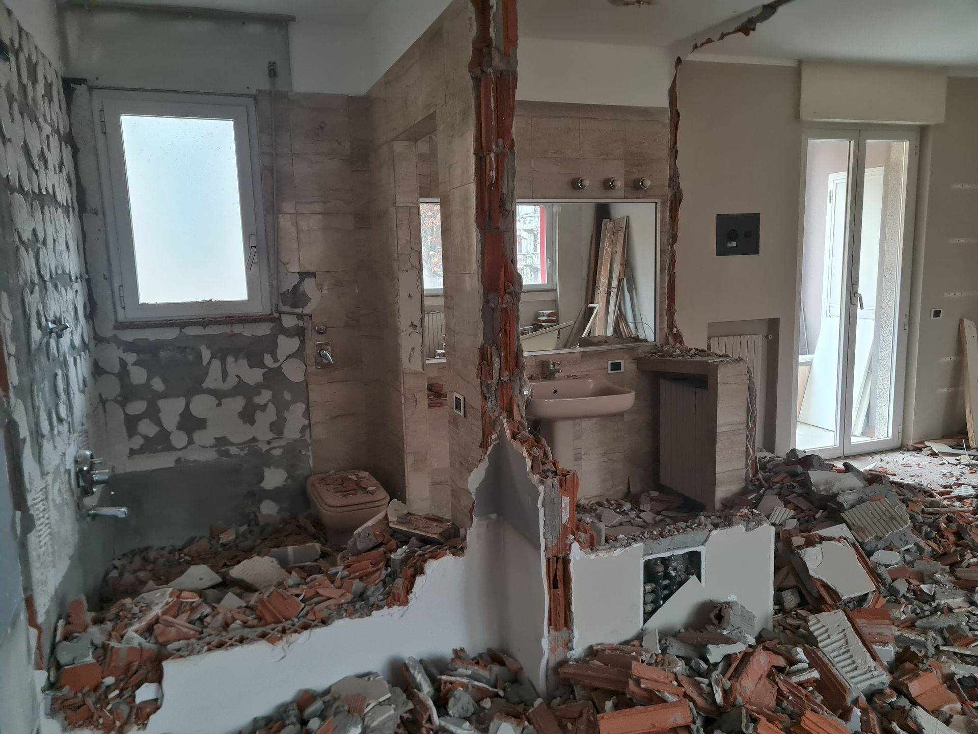 Stanza interna in fase di demolizione, detriti sul pavimento. Pareti parzialmente rotte. Finestra e specchio visibili.