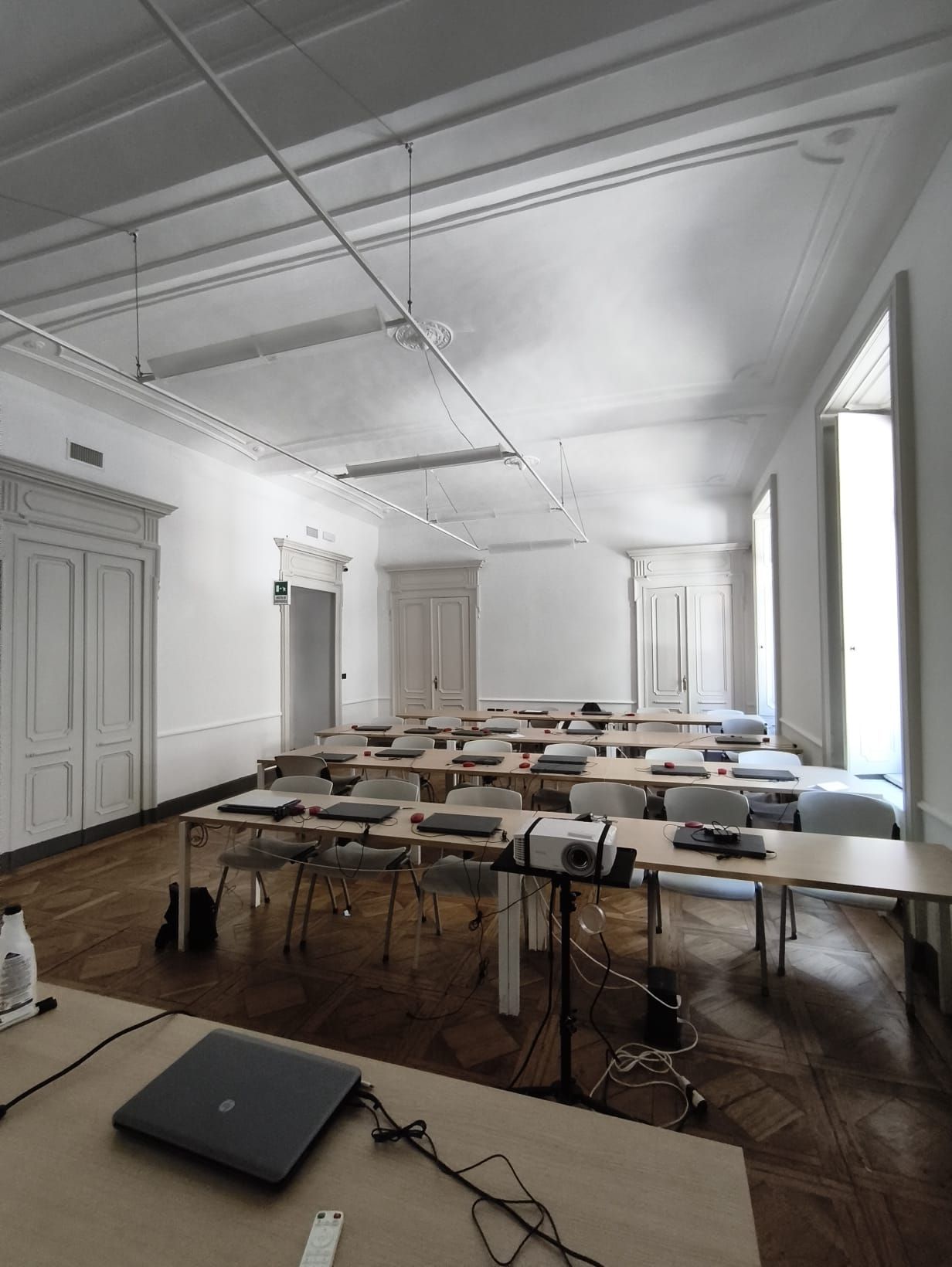 Aula vuota con file di tavoli, computer portatili e un proiettore. Pareti bianche, pavimento in legno, ampie finestre.