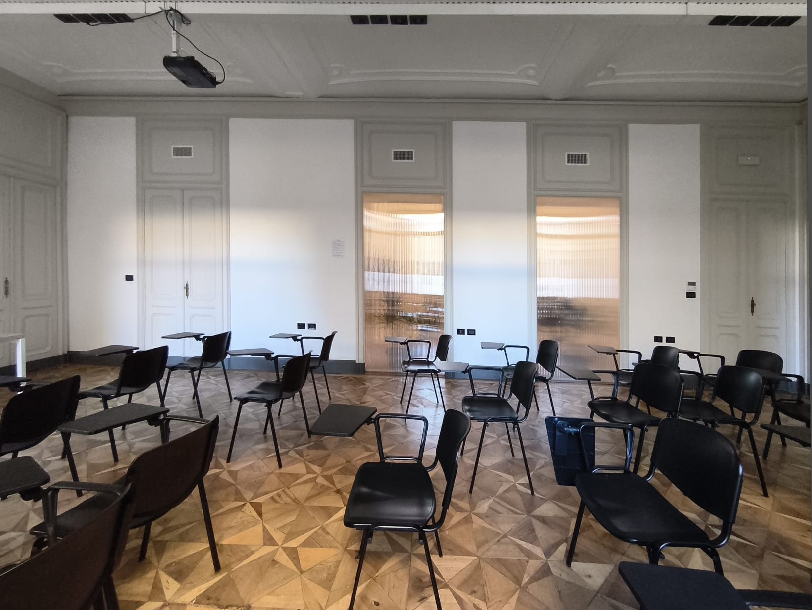 Aula vuota con sedie e tavoli neri su un pavimento di legno, con pareti e finestre bianche.