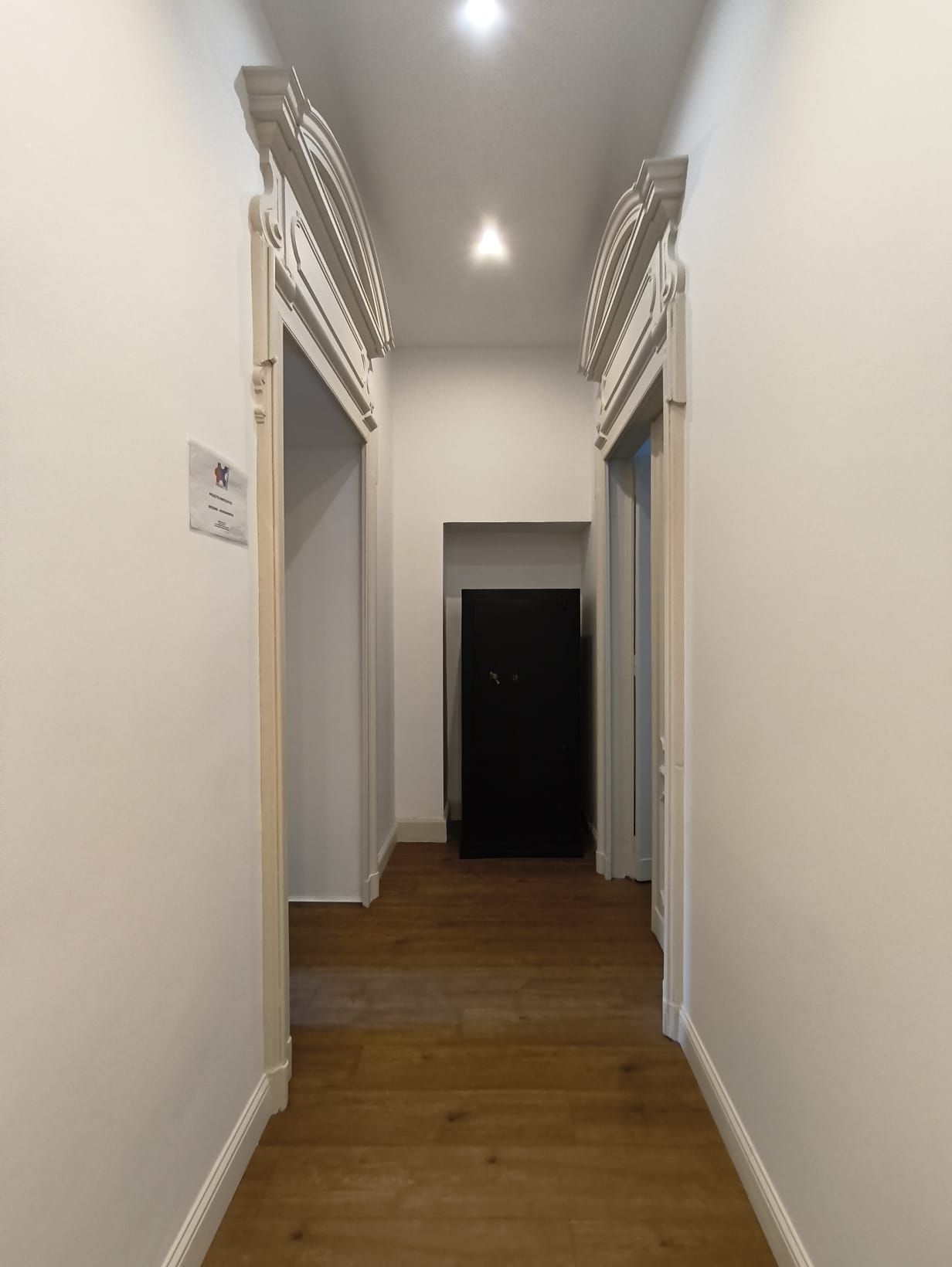 Corridoio con due porte decorate, che conducono a una porta buia in fondo. Pavimento in legno, pareti bianche.
