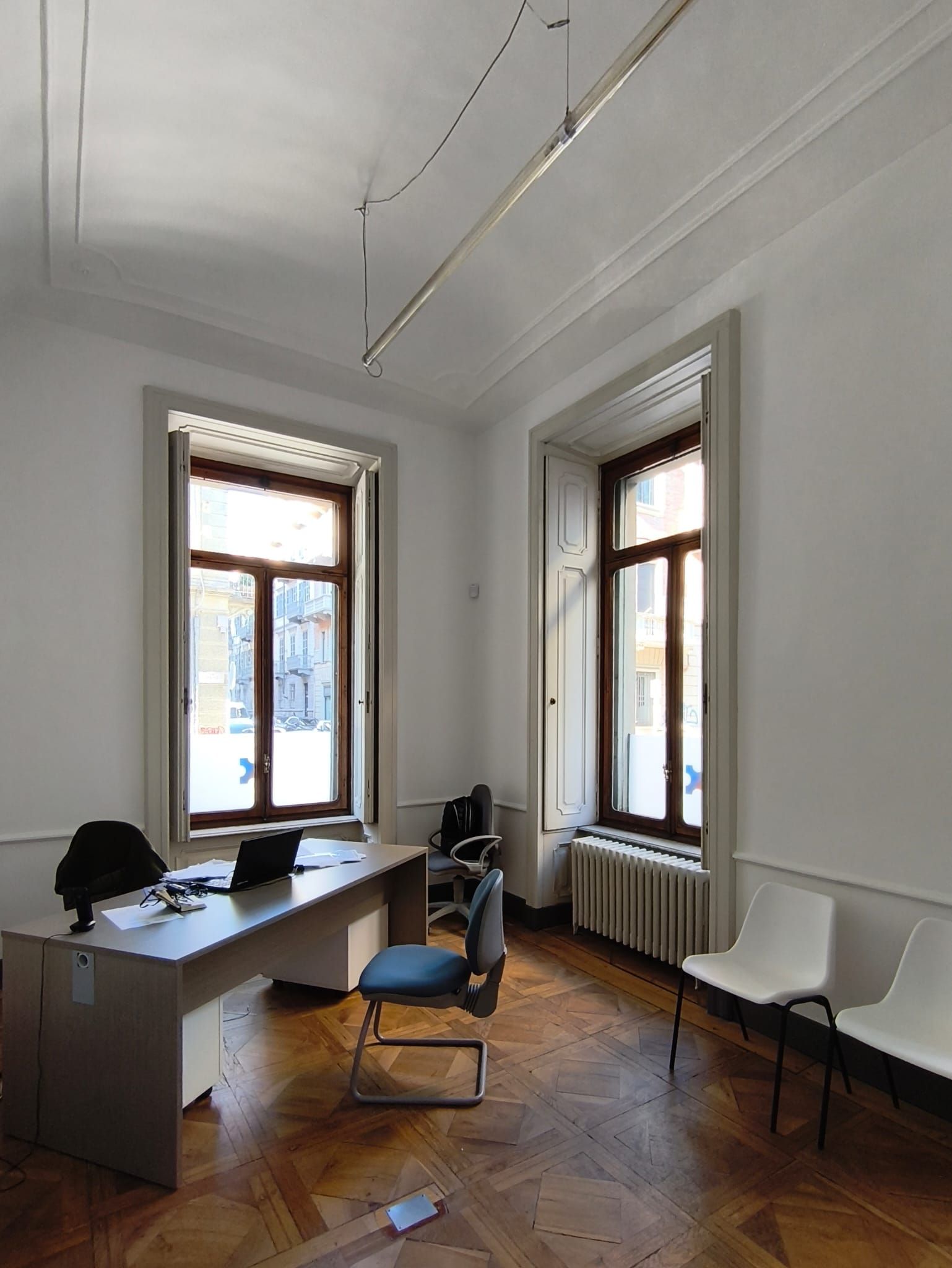 Ufficio con scrivania, finestre, sedie. Pavimento in legno, pareti bianche e termosifone.