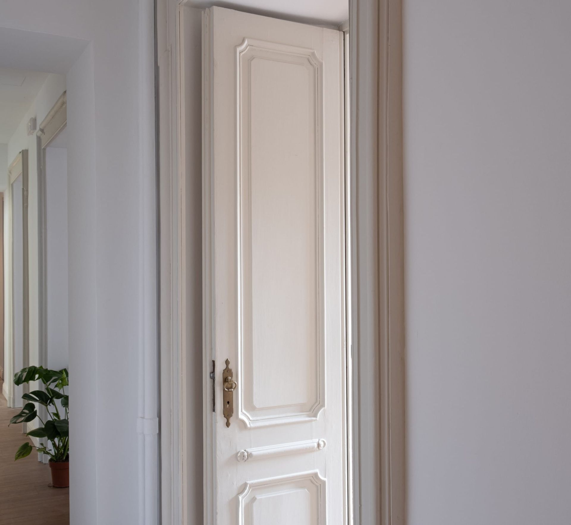 Porta con pannelli bianchi leggermente socchiusa in un corridoio dalle pareti bianche, maniglia in ottone visibile.