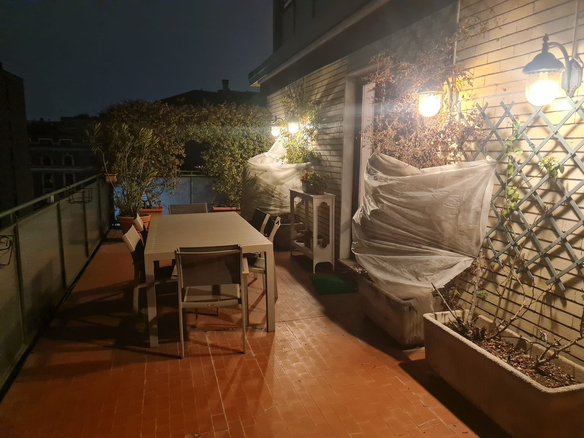Patio esterno di notte, tavolo e sedie, luci a ghirlanda, piante, grata di legno.
