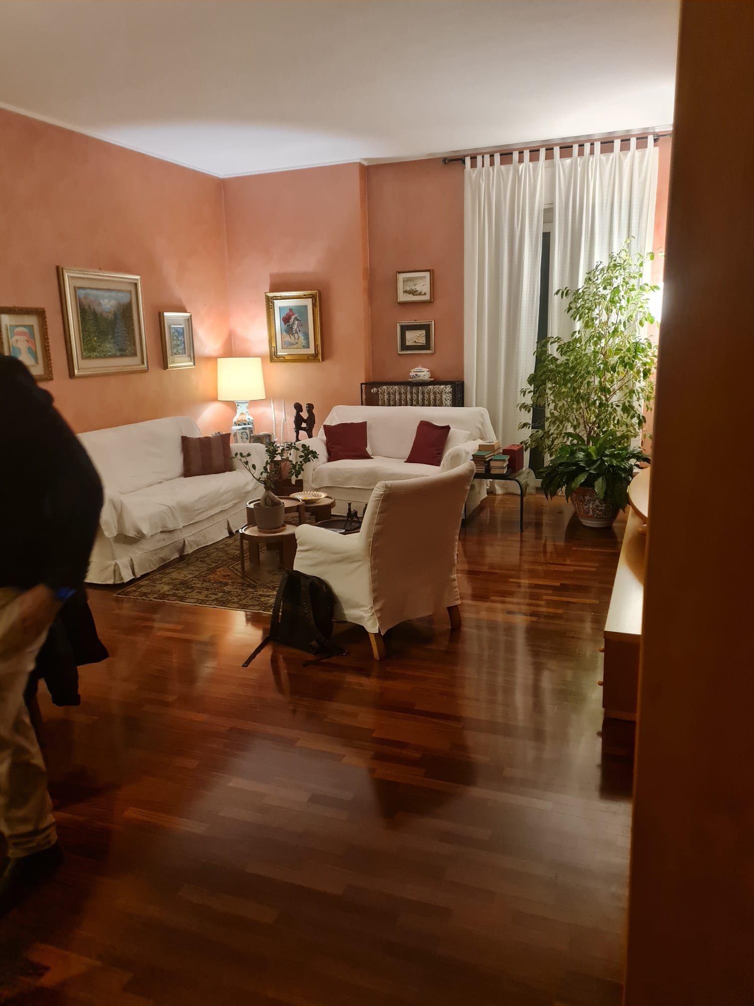 Soggiorno con divani bianchi, pavimento in legno e pareti color pesca; una persona è parzialmente visibile sulla sinistra.