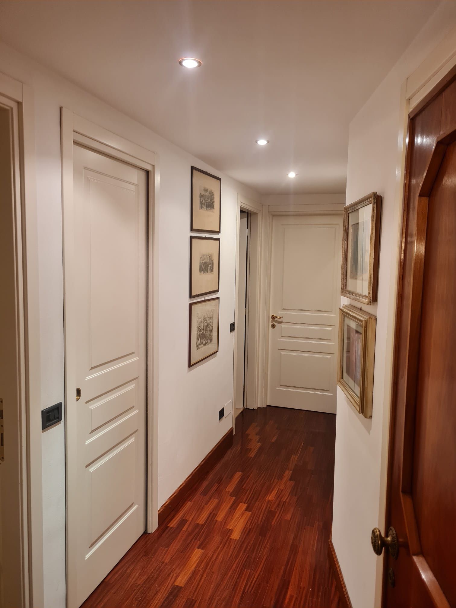 Corridoio stretto con porte bianche, quadri incorniciati e pavimento in legno scuro.