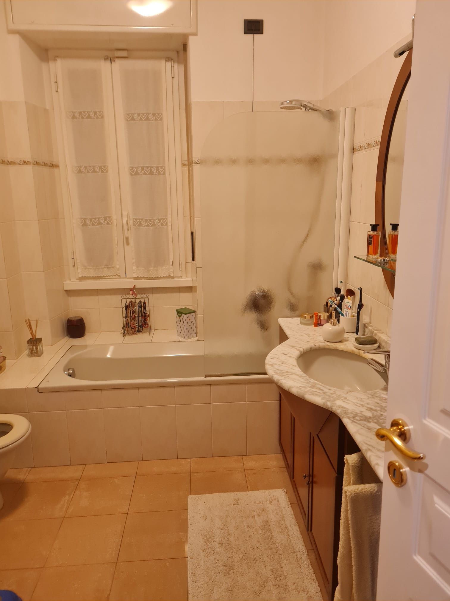 Bagno con vasca, lavandino, WC e finestra. Piastrelle beige e luci calde.