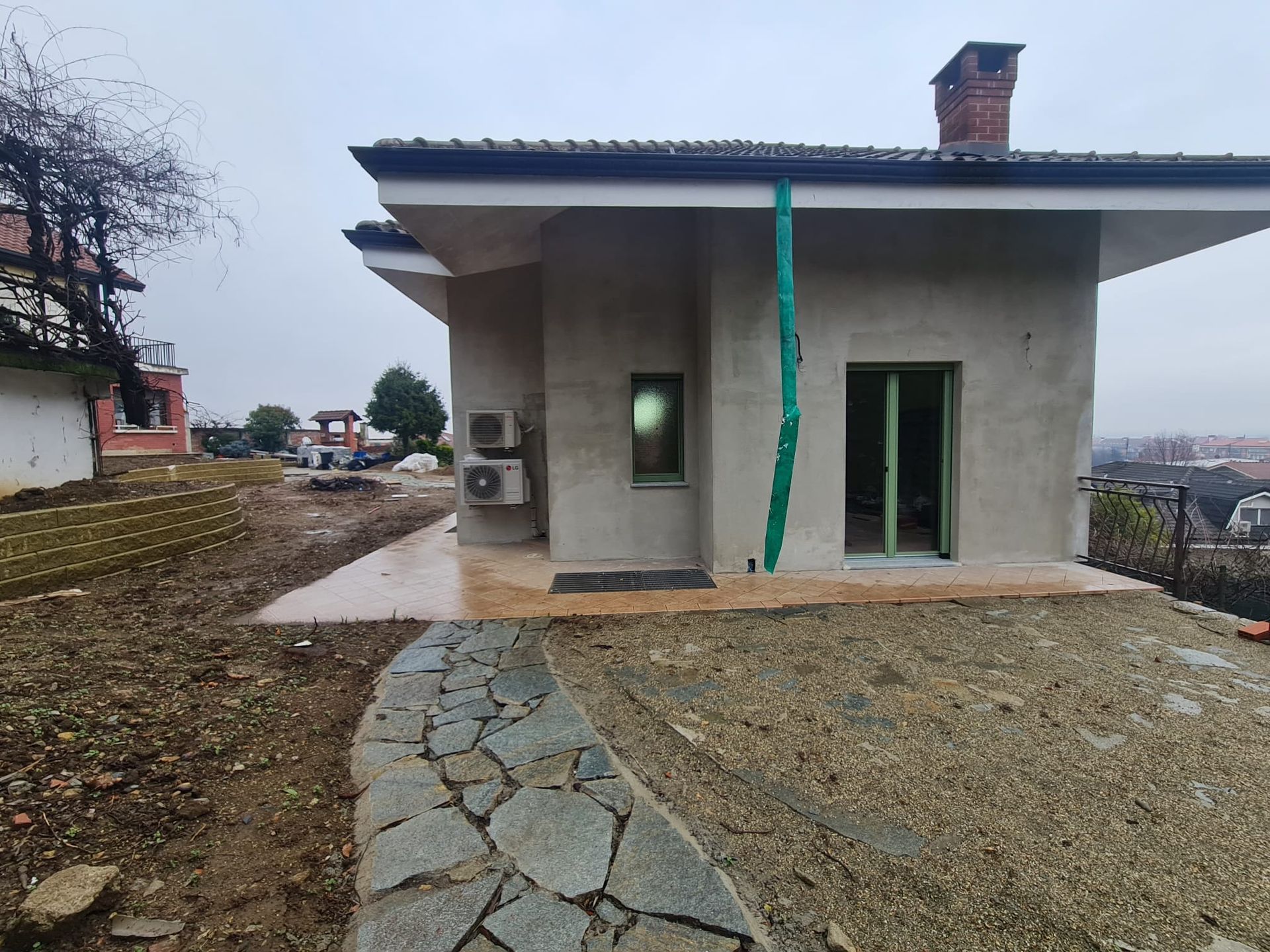 Piccola casa incompiuta con sentiero in pietra e tubo verde in una giornata nuvolosa.