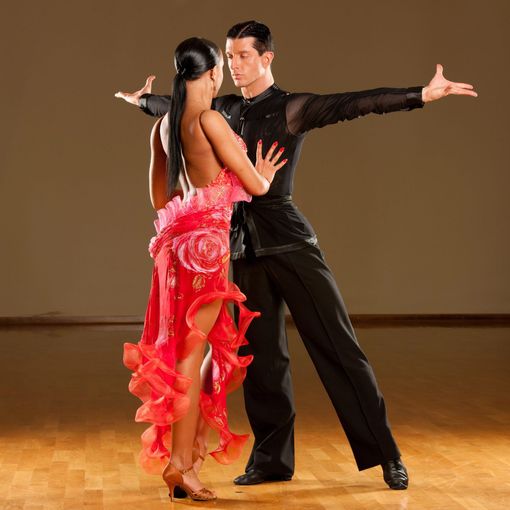 Couple performing ballroom dance on a wooden floor, woman in red dress, man in black suit, arms outstretched.