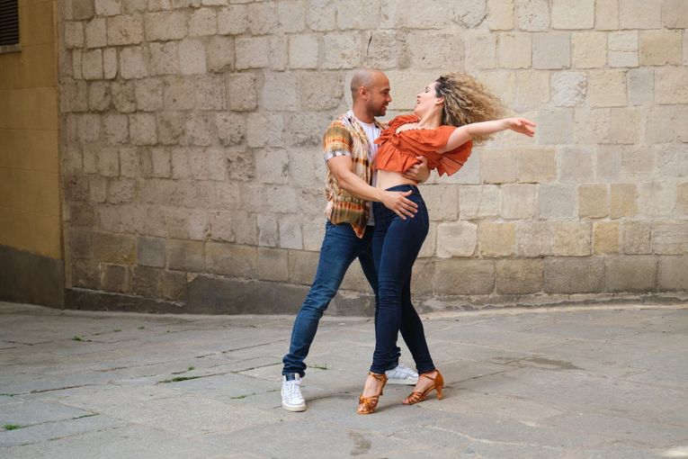 Couple dancing salsa in a stone-walled alleyway; woman leans back, hair flying; man holds her waist, looking at her.