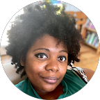 A person with curly dark hair looks toward the camera with a soft smile, set against a blurred indoor background.