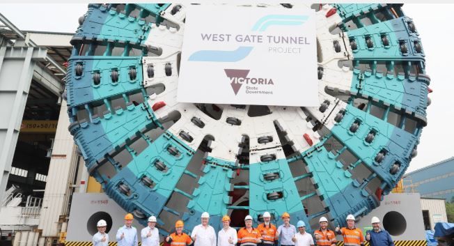 A group of people are standing in front of a large machine that says west gate tunnel.
