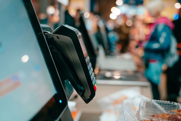 Close-up of a payment terminal beside a store checkout screen with a blurred shopper in the background