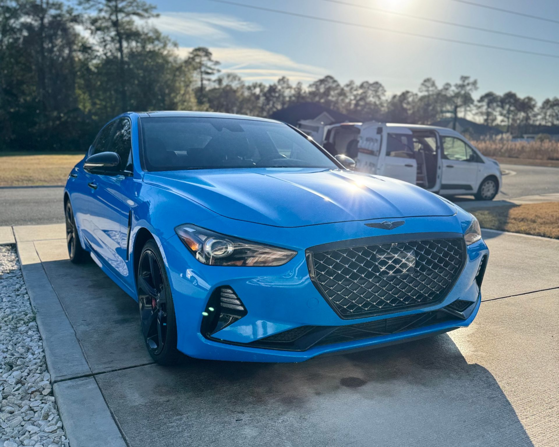 Blue Genesis G70 sedan parked on a driveway in front of a white van, sunny day.