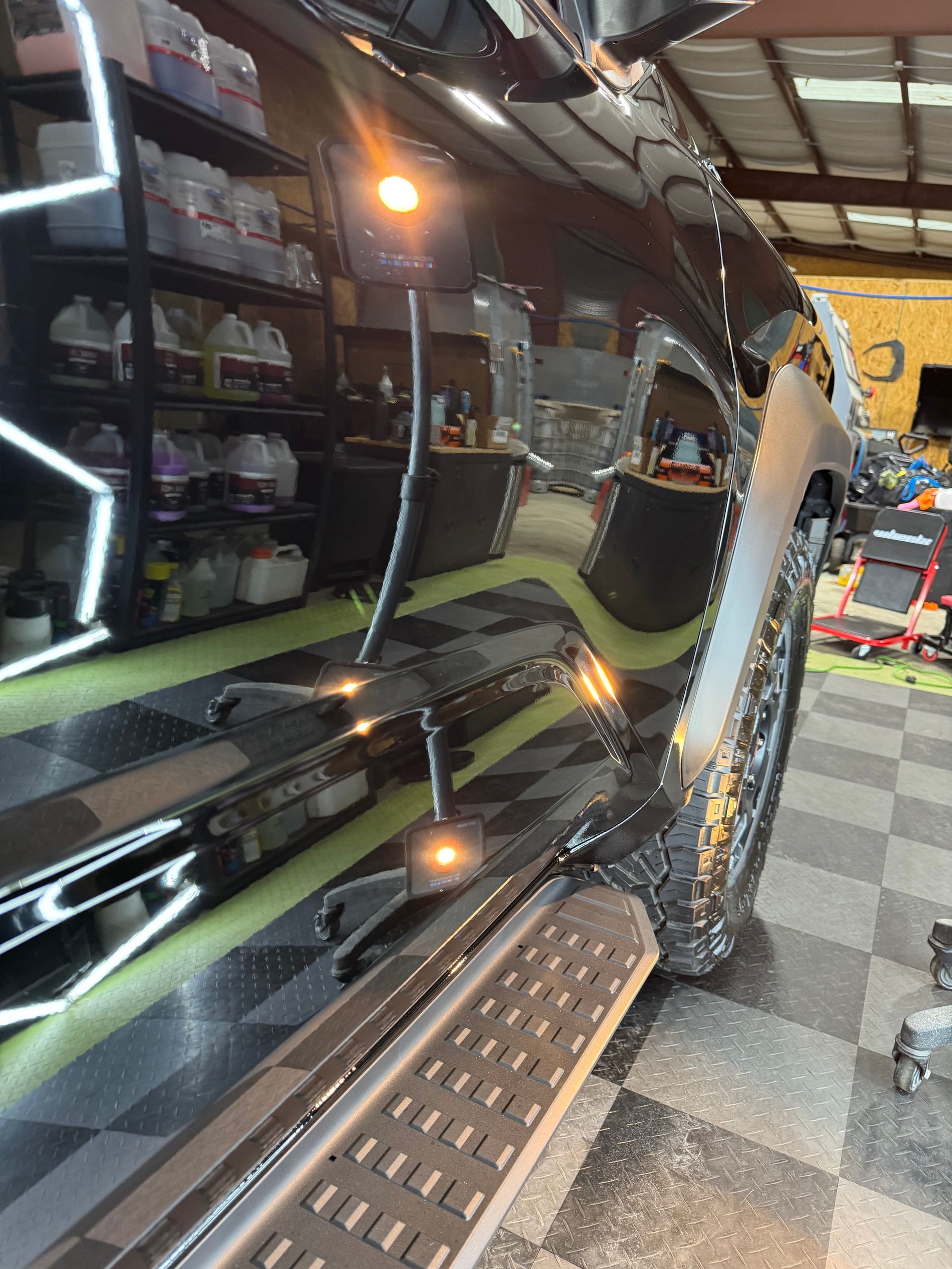 Black vehicle's glossy side reflecting shop lights and the tiled floor. Running board visible.