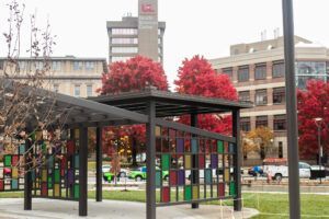 A modern pavilion with colorful stained-glass panels, set in a grassy park with bright red autumn trees and tall buildings.