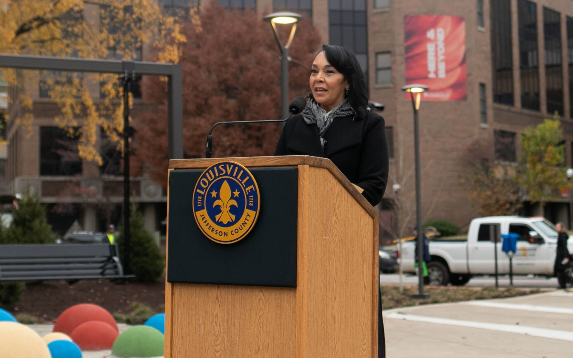 A speaker stands at a podium with a Louisville seal outdoors, addressing an audience in front of a brick building.