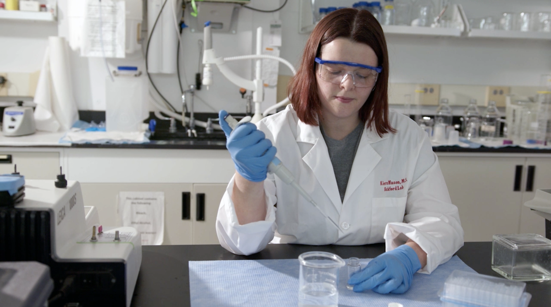 A researcher in a white lab coat and safety goggles uses a pipette to work with samples on a laboratory bench.