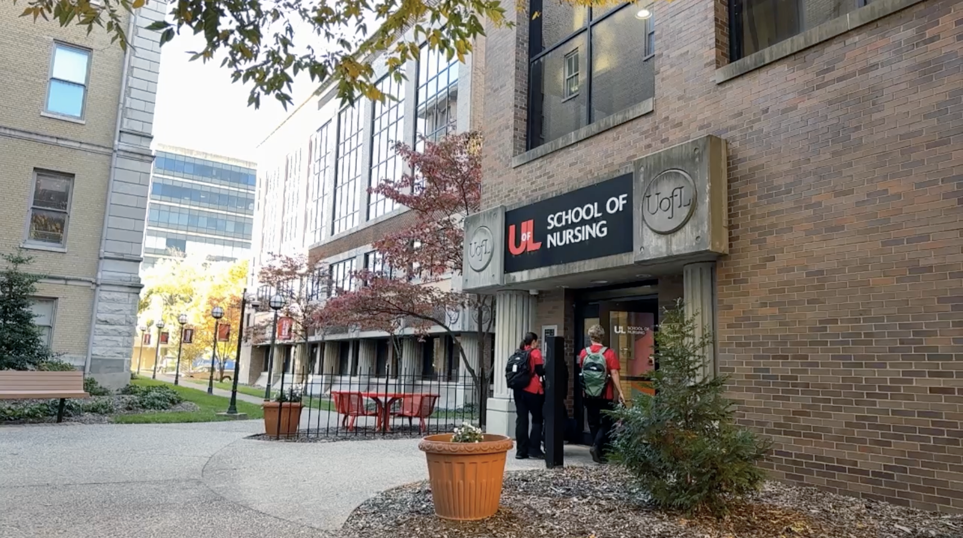 People entering the University of Louisville School of Nursing building on a sunny day with trees and outdoor seating.