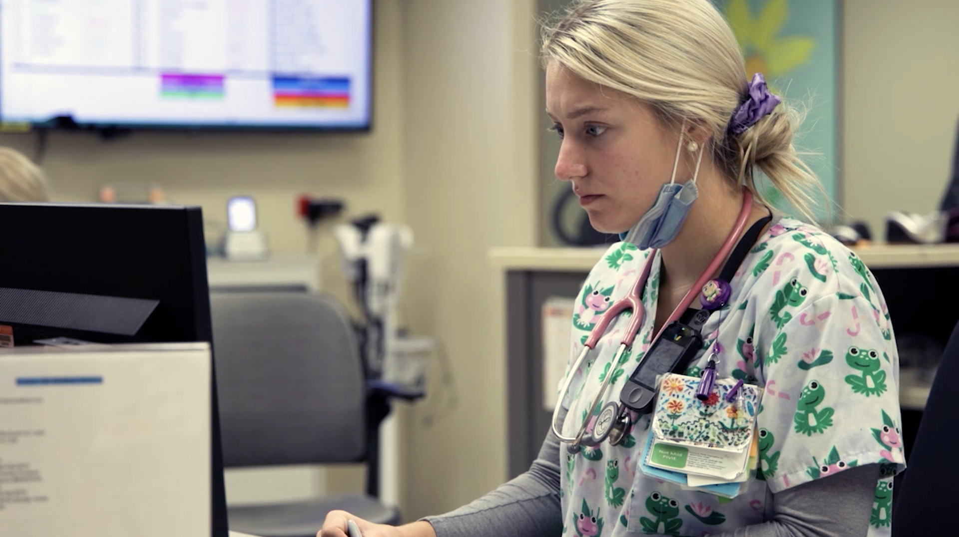 A healthcare worker wearing a patterned scrub top and a stethoscope, working at a computer in a clinical office.