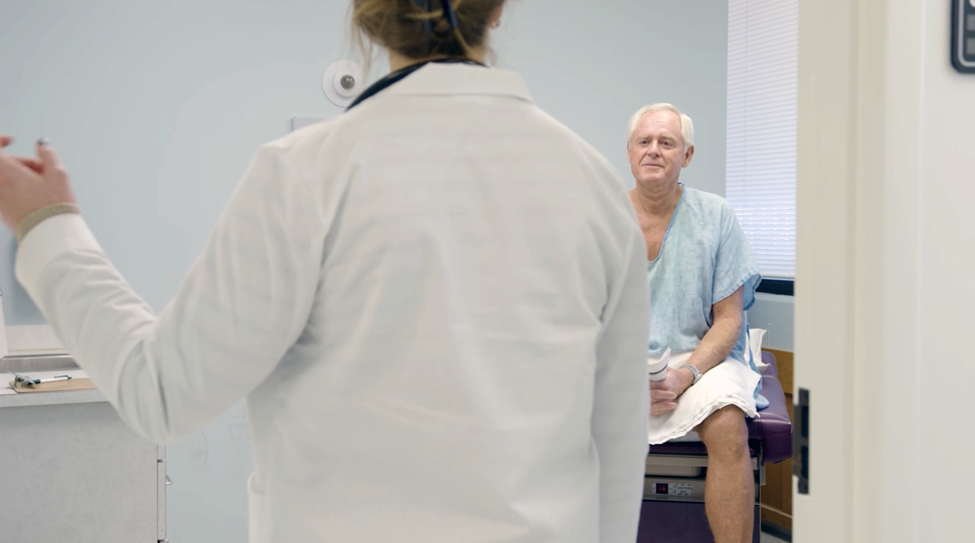 A healthcare professional in a white coat gestures while speaking to a patient sitting on an exam table in a clinic.