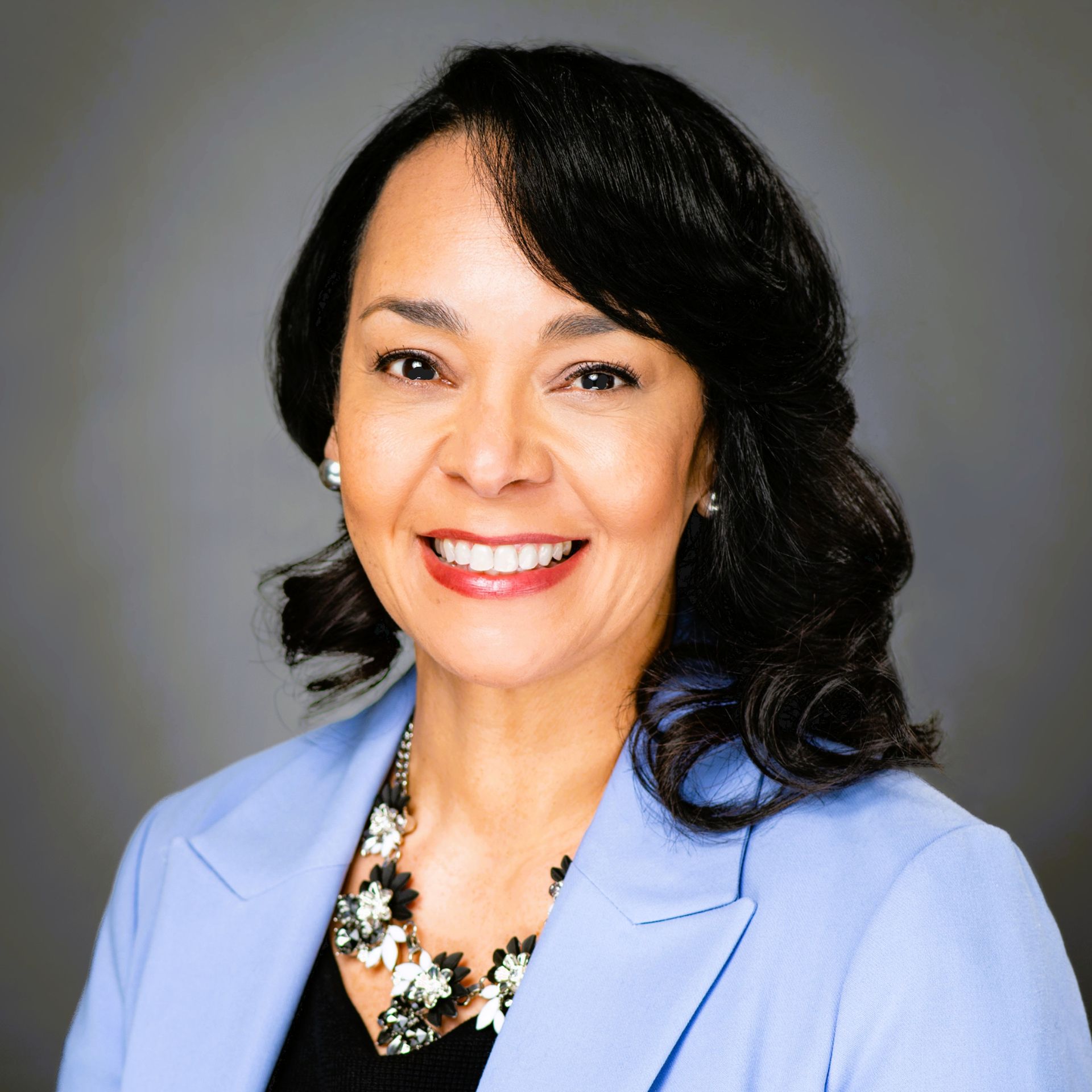 A smiling professional in a light blue blazer and statement necklace against a grey background.