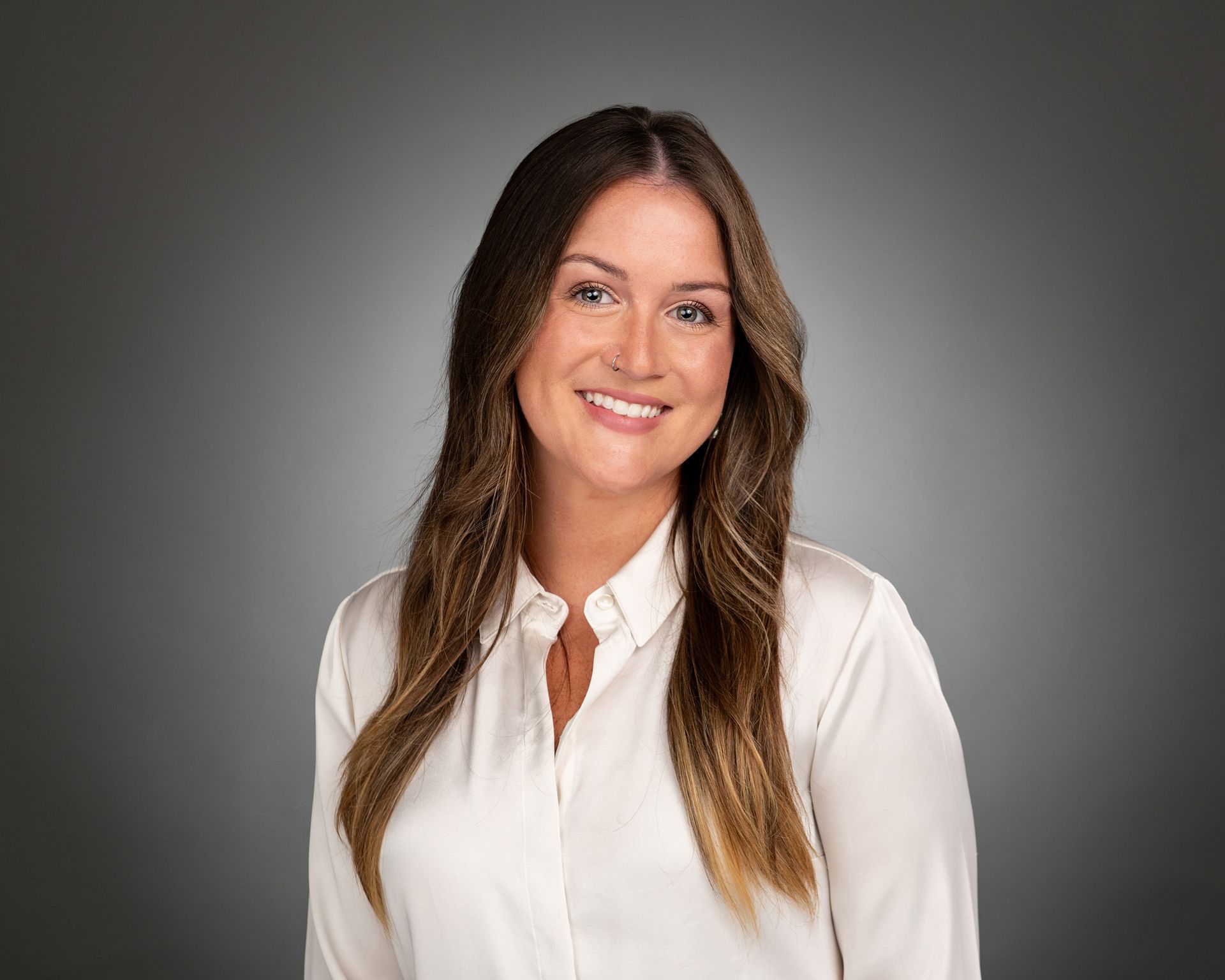A professional headshot of a smiling person with long brown hair wearing a white collared shirt against a gray background.
