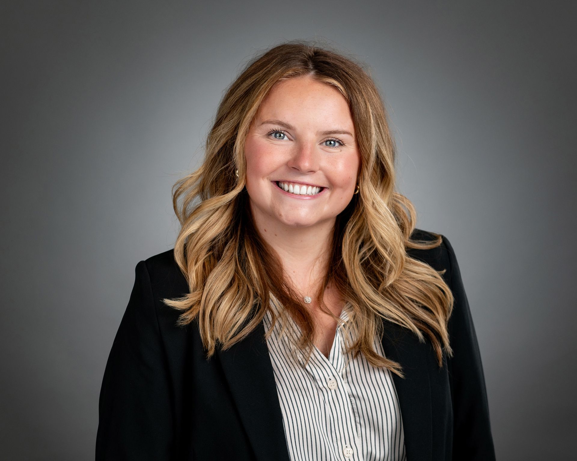 A person with long, wavy blonde hair smiling at the camera, wearing a black blazer over a pinstriped shirt.