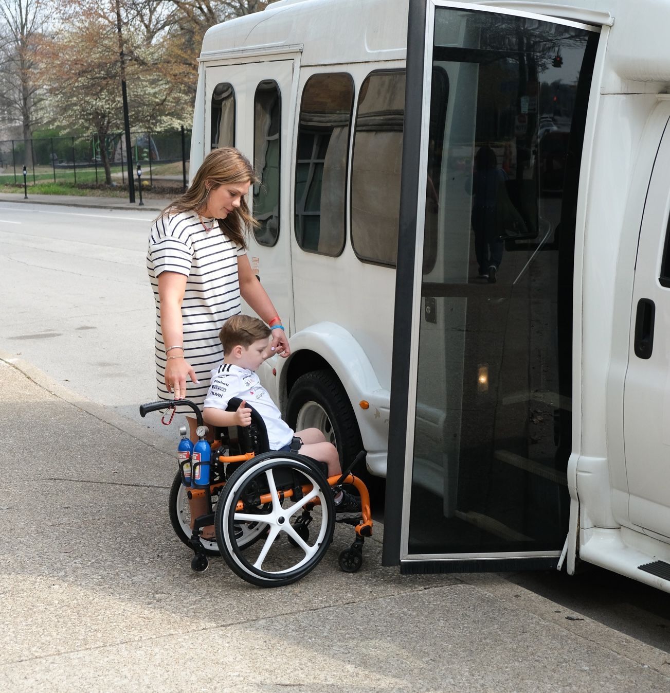 Person with striped shirt helping a child in a wheelchair into a white shuttle bus. 