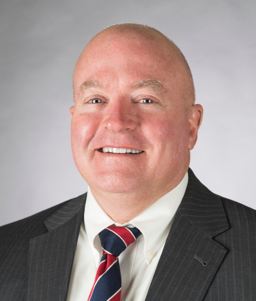 A man smiling, wearing a dark pinstripe suit, white shirt, and a red and blue striped tie against a gray background.