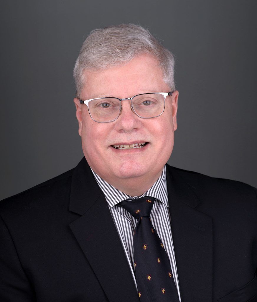 A professional headshot of a person wearing glasses, a dark suit, and a striped shirt with a dark patterned tie.