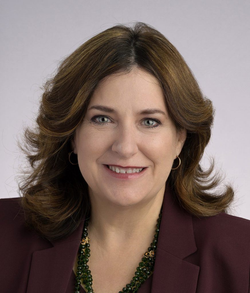 A smiling woman with shoulder-length brown hair, wearing a dark blazer and a green beaded necklace.