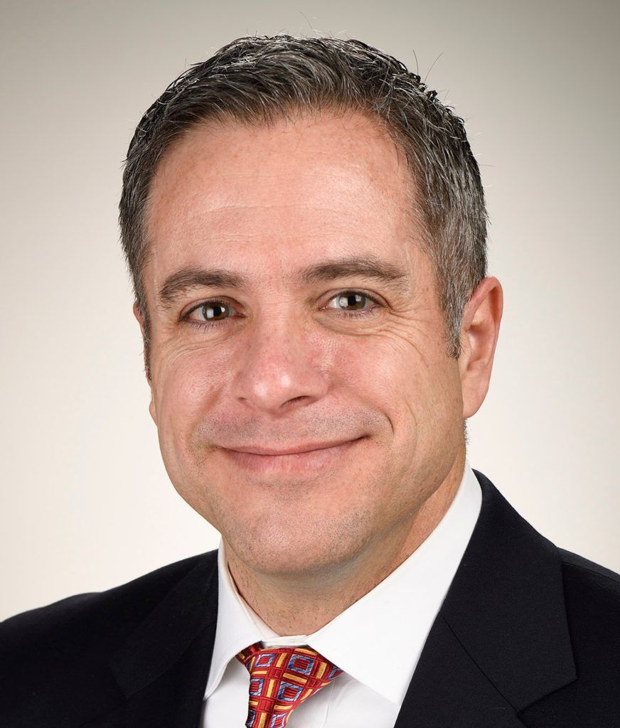 A professional headshot of a man with short brown hair, wearing a dark suit, white shirt, and a red patterned tie.