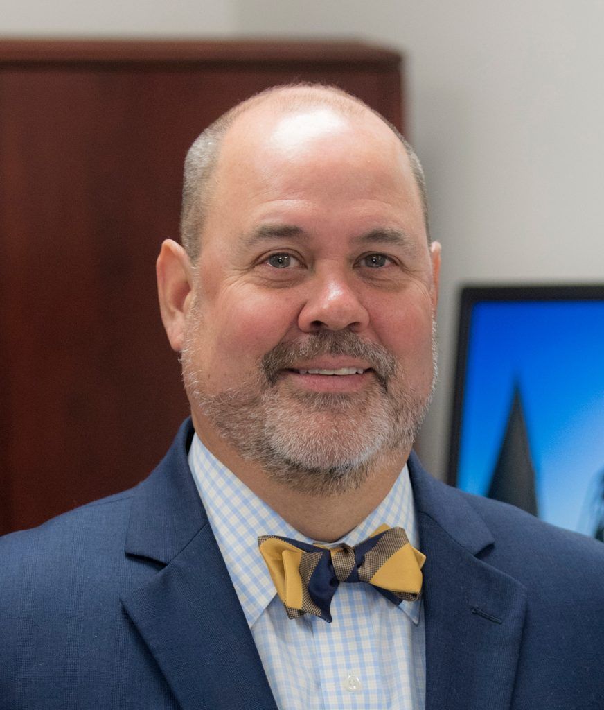 A man with a short beard wearing a blue suit, light-checked shirt, and a yellow and navy bowtie smiles at the camera.