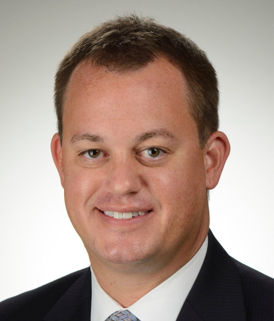 A professional headshot of a smiling man in a suit and tie against a plain light gray background.