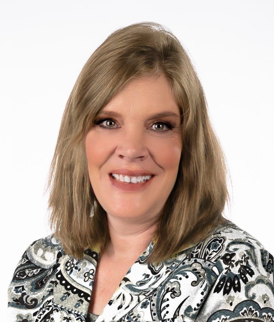 A person with shoulder-length light brown hair smiling at the camera, wearing a black and white paisley-patterned shirt.
