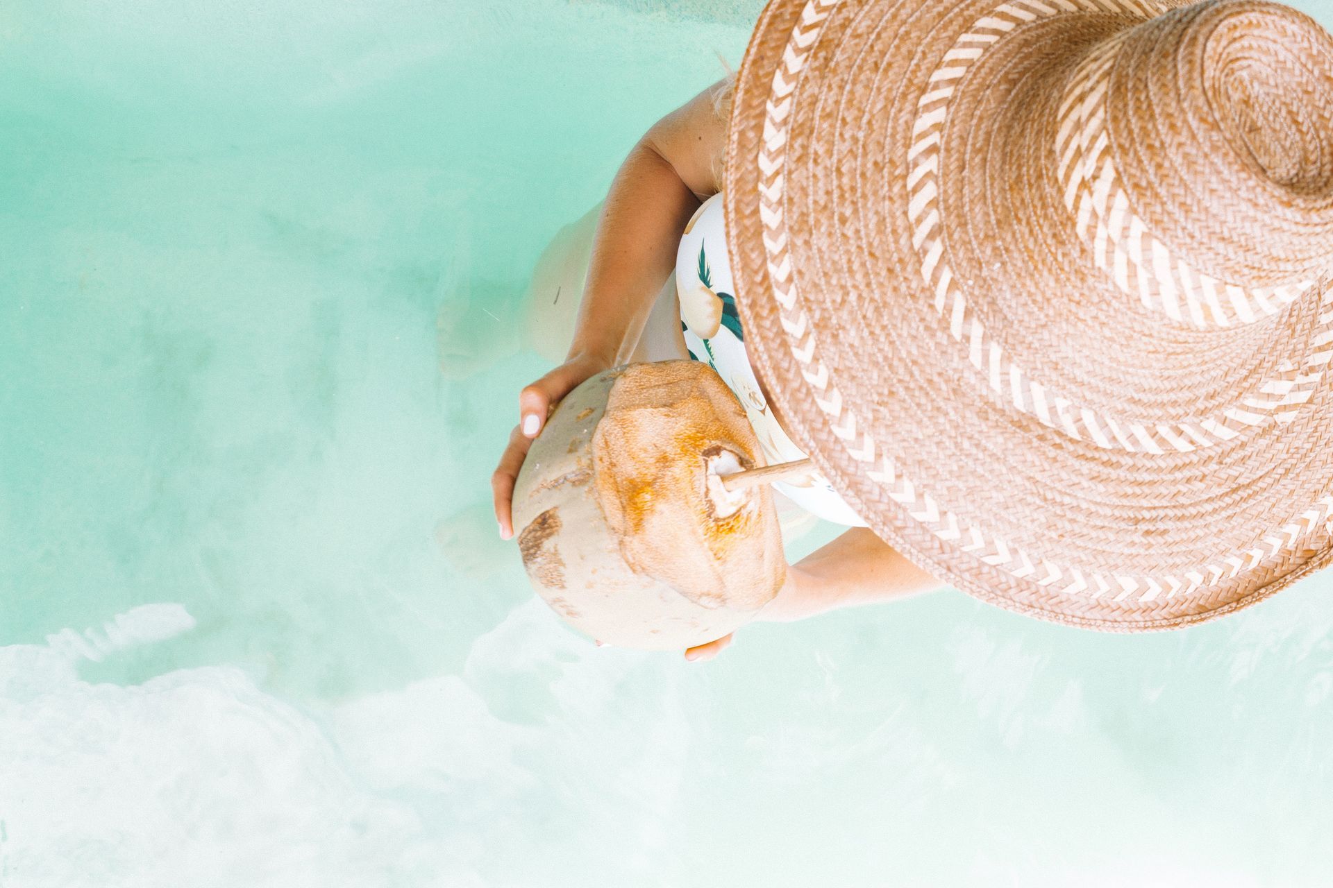A woman wearing a straw hat is drinking from a coconut in a pool.