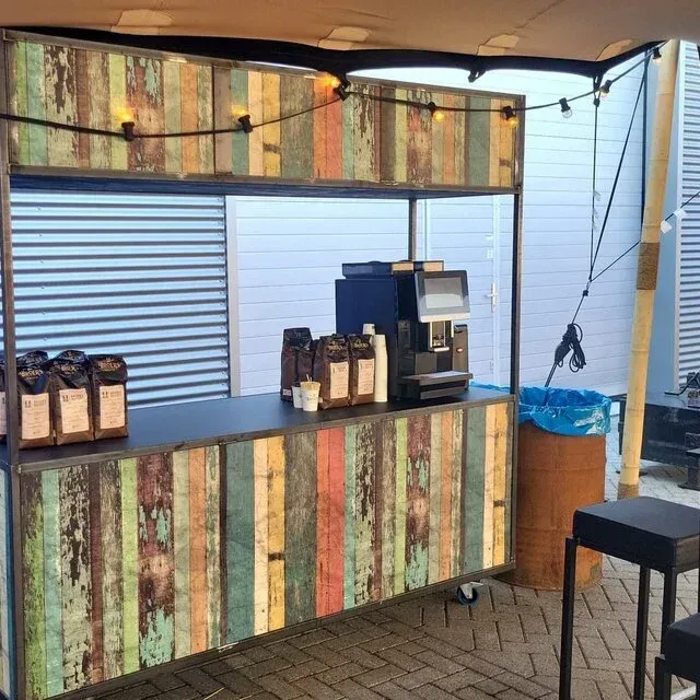 Koffiekar met verweerde houten panelen, onder een luifel. Koffiezetapparaat, bonen en prullenbak zichtbaar.