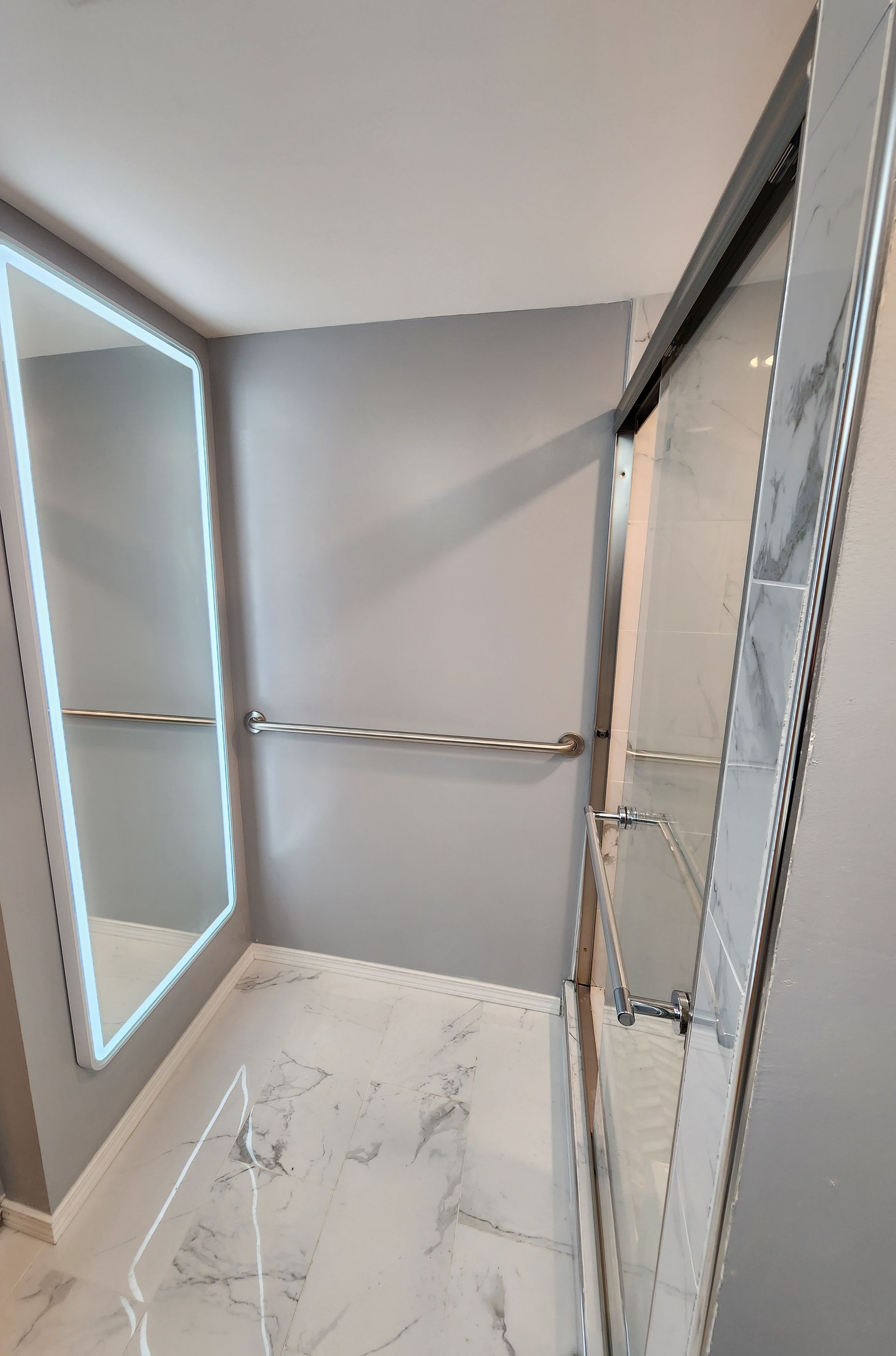 Modern bathroom with gray walls, marble floor, and a backlit mirror beside a glass shower enclosure