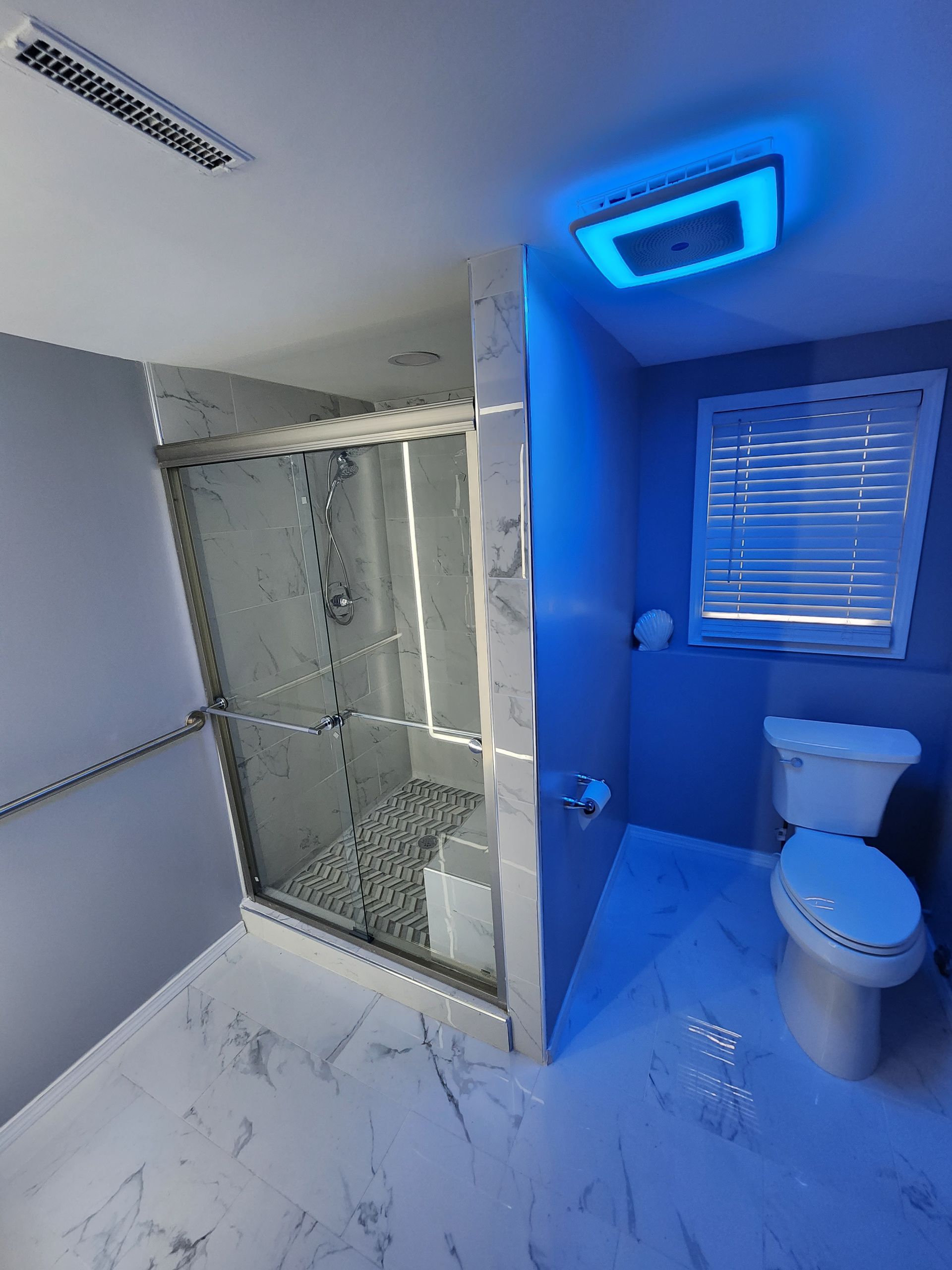 Blue-lit bathroom with glass shower, toilet, window blinds, and marble-look floor