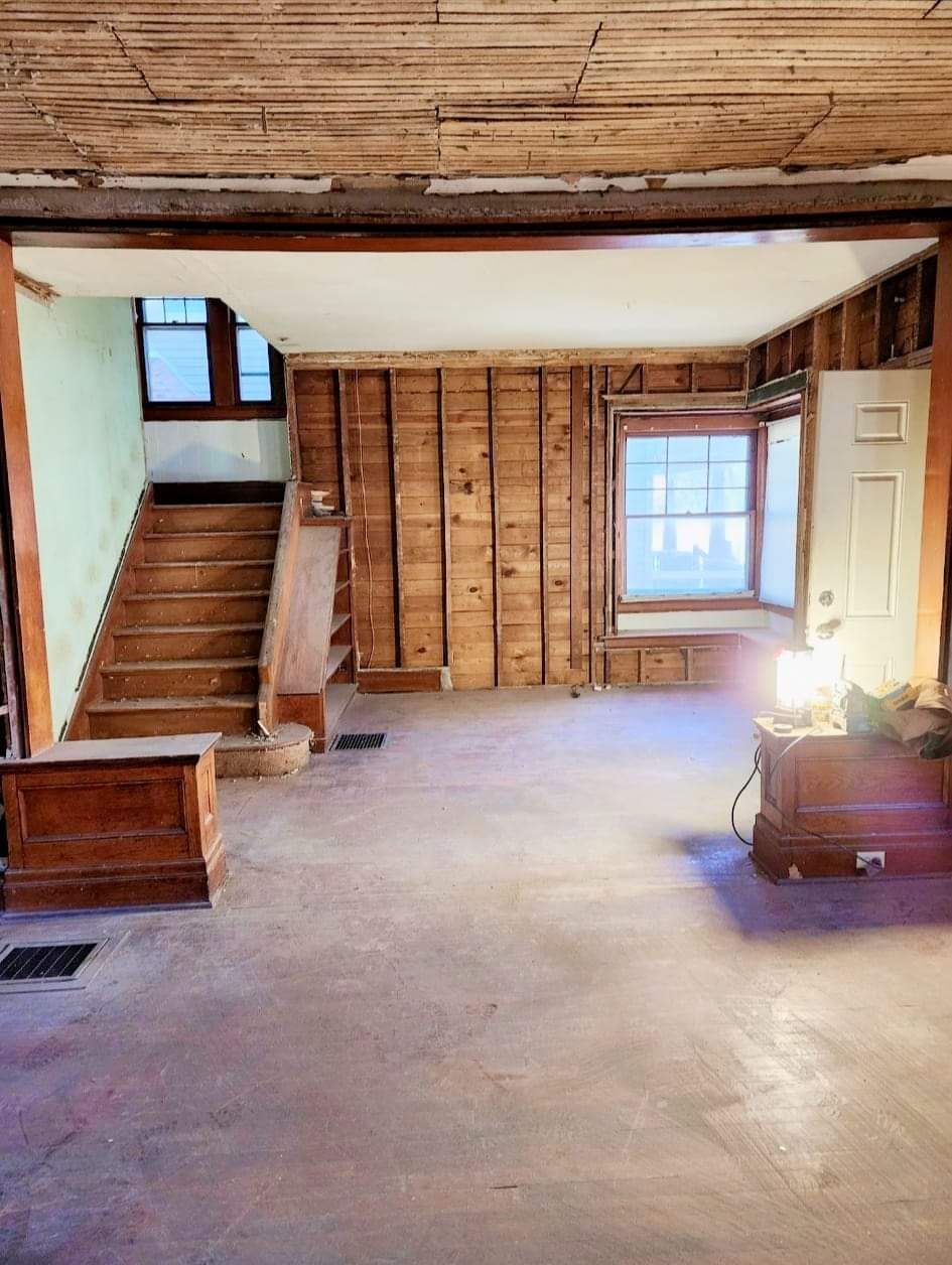 Empty unfinished room with exposed wooden beams, stairs on the left, and a window on the right.
