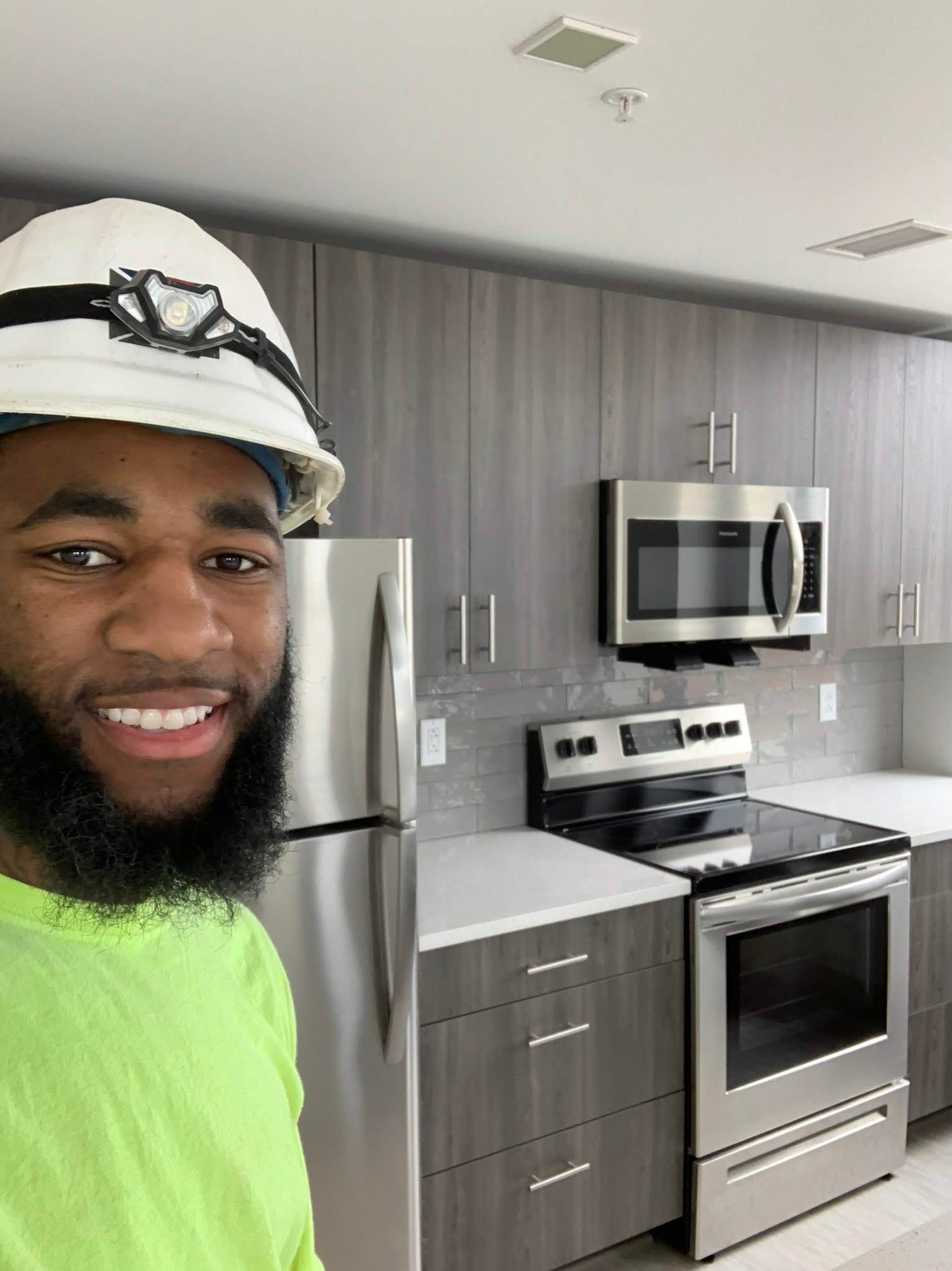 Smiling person in a hard hat standing in a modern kitchen with stainless steel appliances and gray cabinets