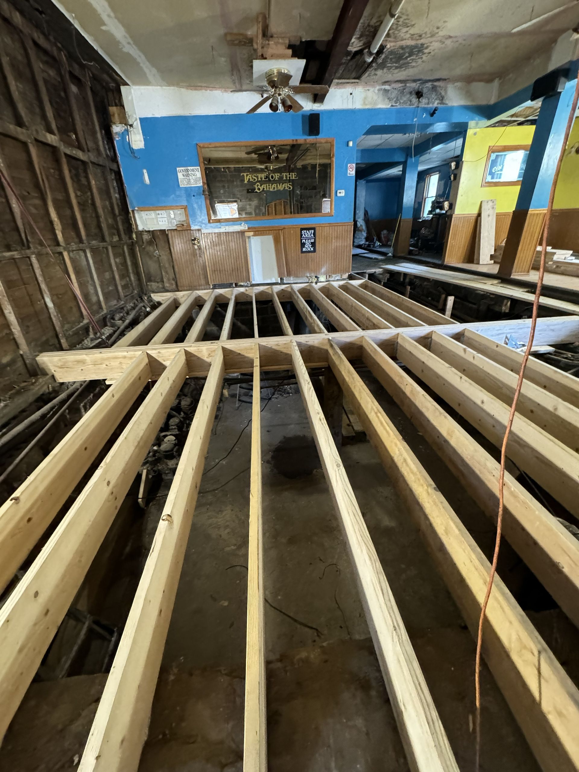 Partially built wooden floor framing inside a damaged room with blue walls and exposed ceiling beams