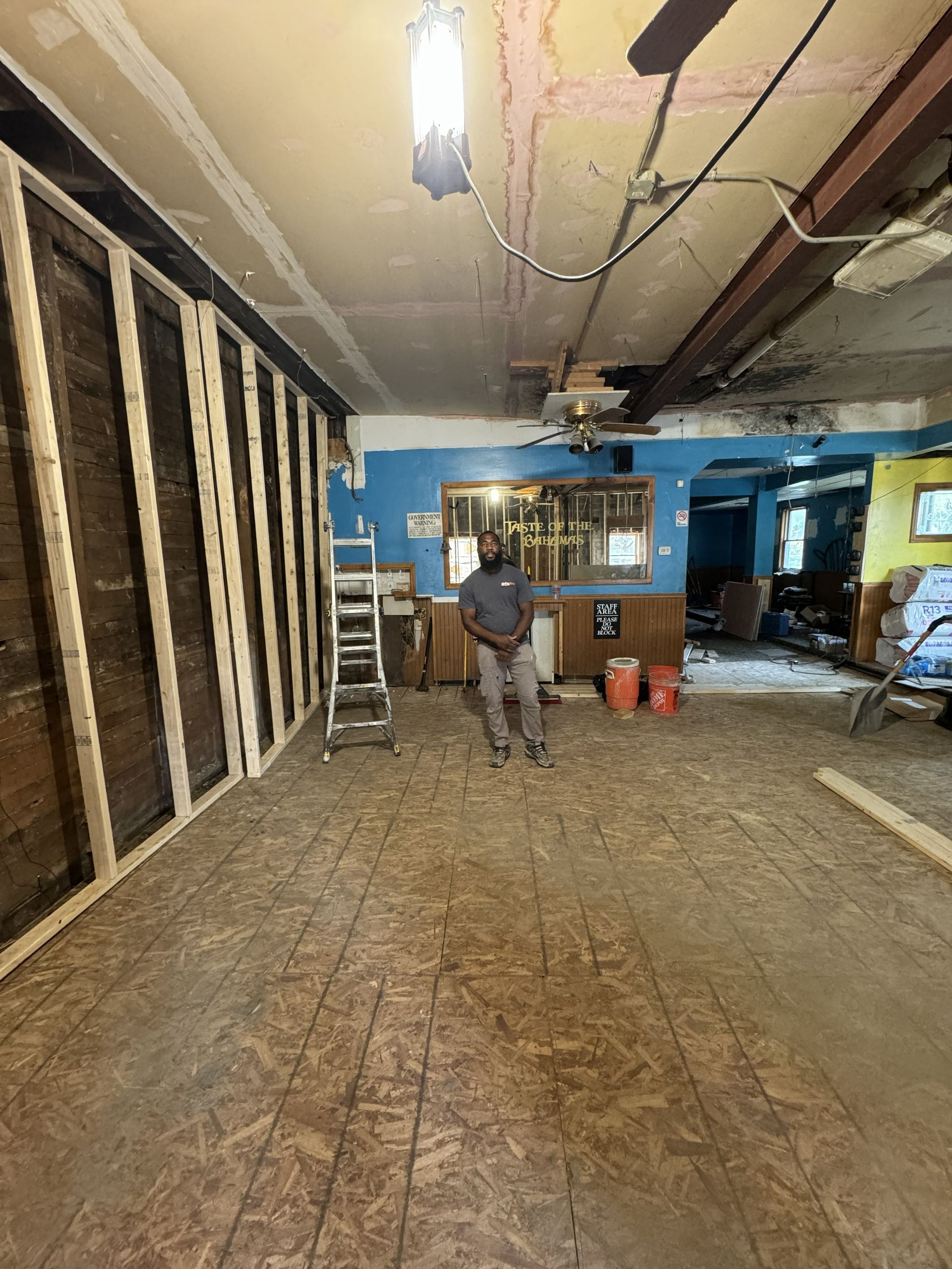 Renovation site with exposed wall studs, ladder, tools, and two workers in a partially stripped room.