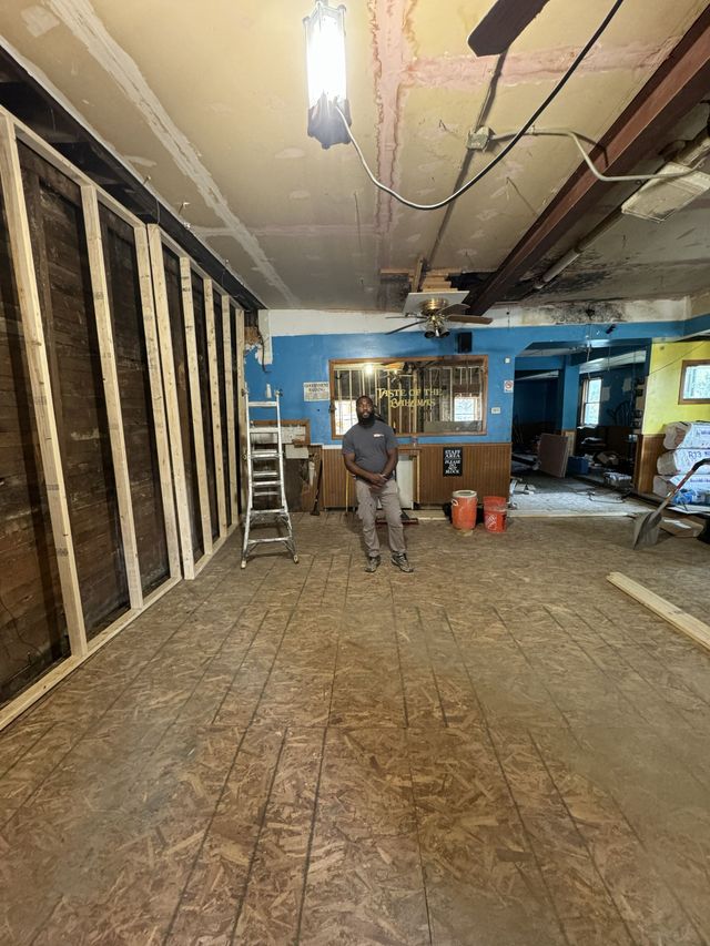 Renovation site with exposed wall studs, ladder, tools, and two workers in a partially stripped room.