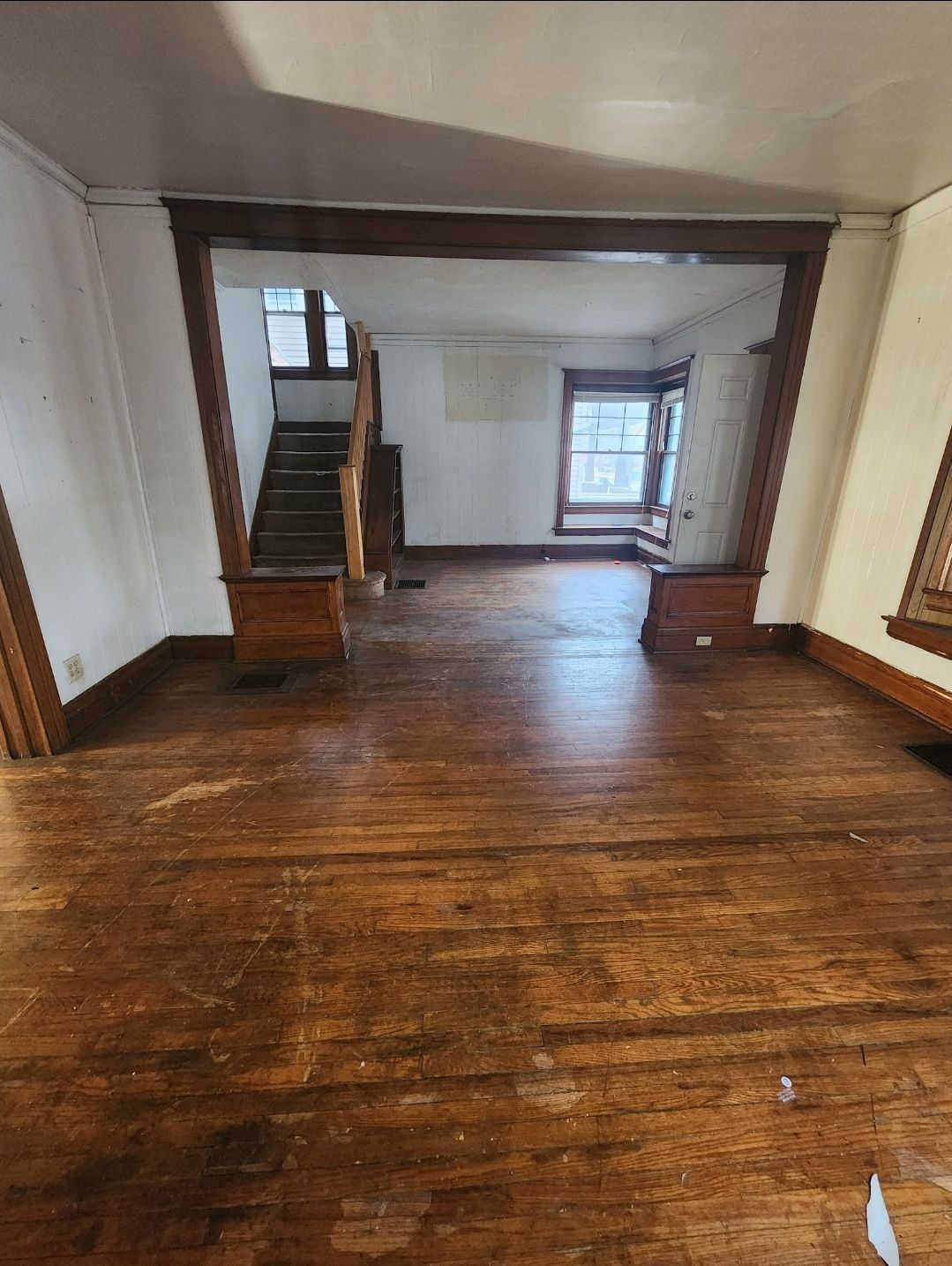 Empty room with worn wooden floors, staircase at left, and a bright window at the back.