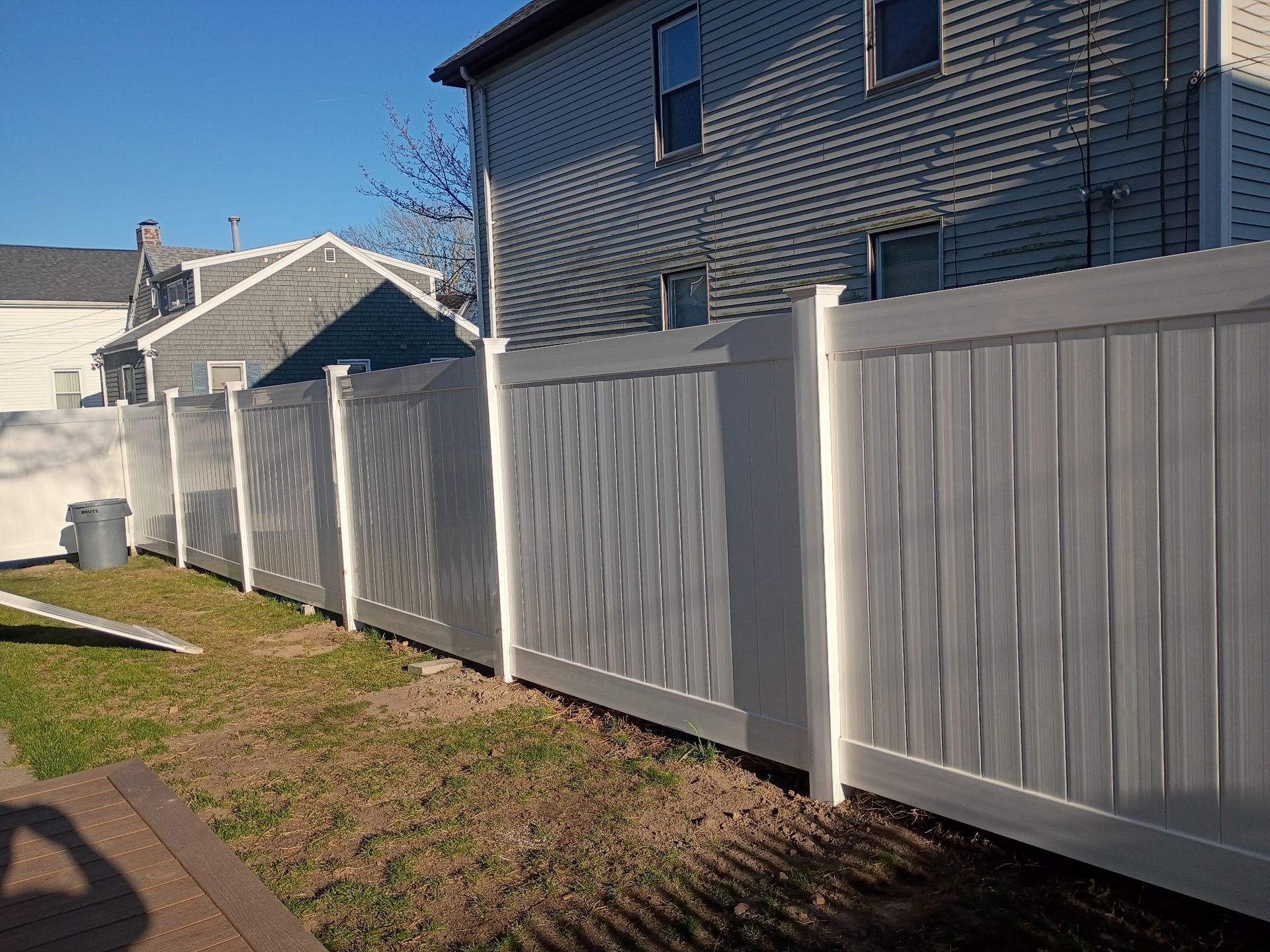 White vinyl privacy fence along a grassy yard beside suburban houses under a clear blue sky