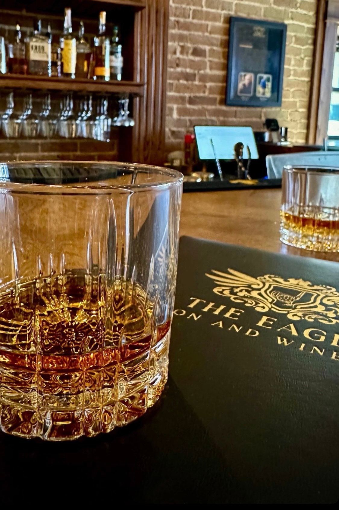 Whiskey in a cut glass on a bar with a menu, brick wall, and bottles.