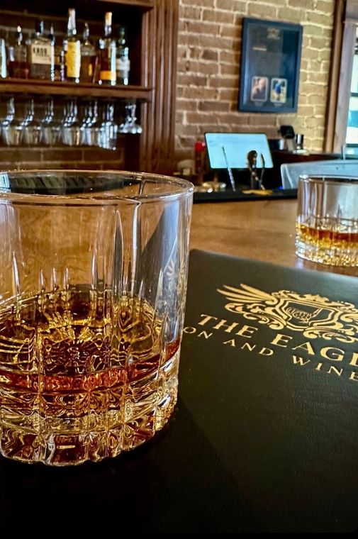 Whiskey in a cut glass on a bar with a menu, brick wall, and bottles.